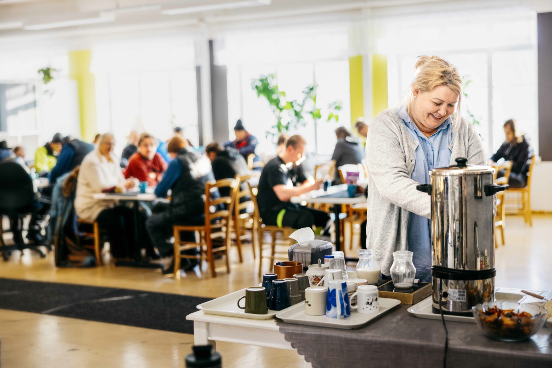 Ihmisiä kahvipöytien äärellä. Etualalla keski-ikäinen nainen ottaa kahvia termostaatista.